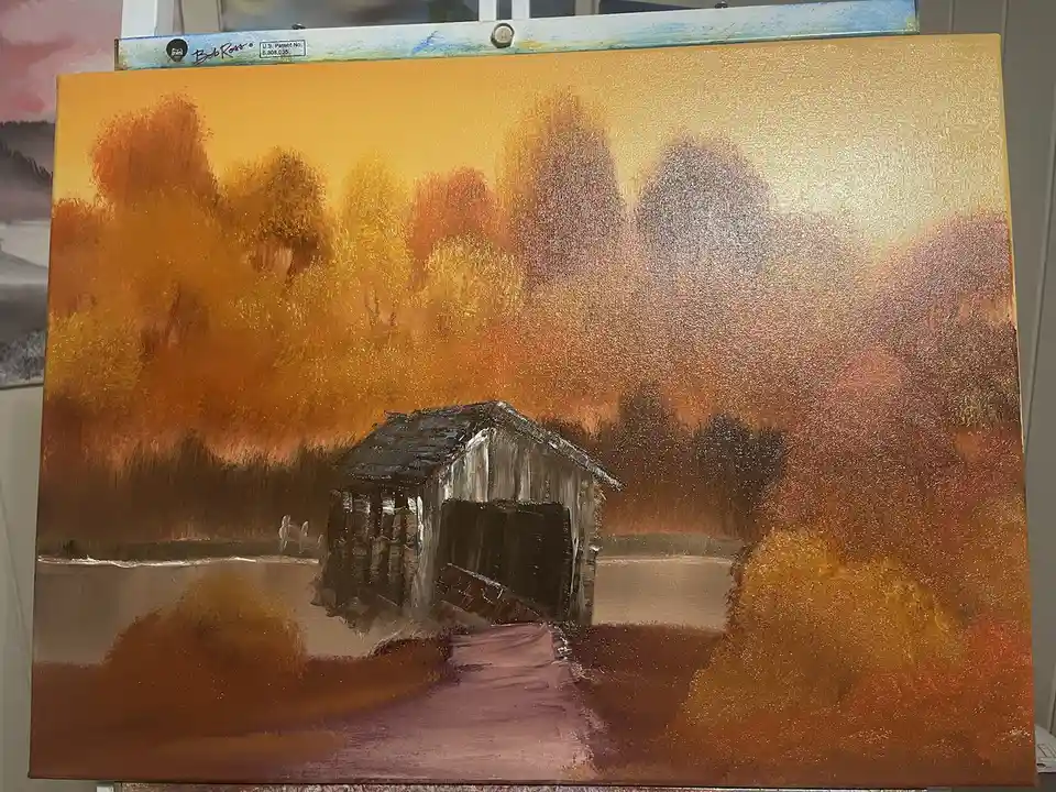 A painting on canvas depicts a rustic wooden bridge over water, surrounded by autumnal trees in orange and brown hues under…