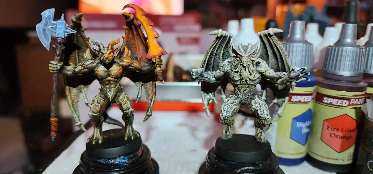 Two painted fantasy miniatures stand side-by-side on black bases; the left figure holds an axe and has fiery orange wings…