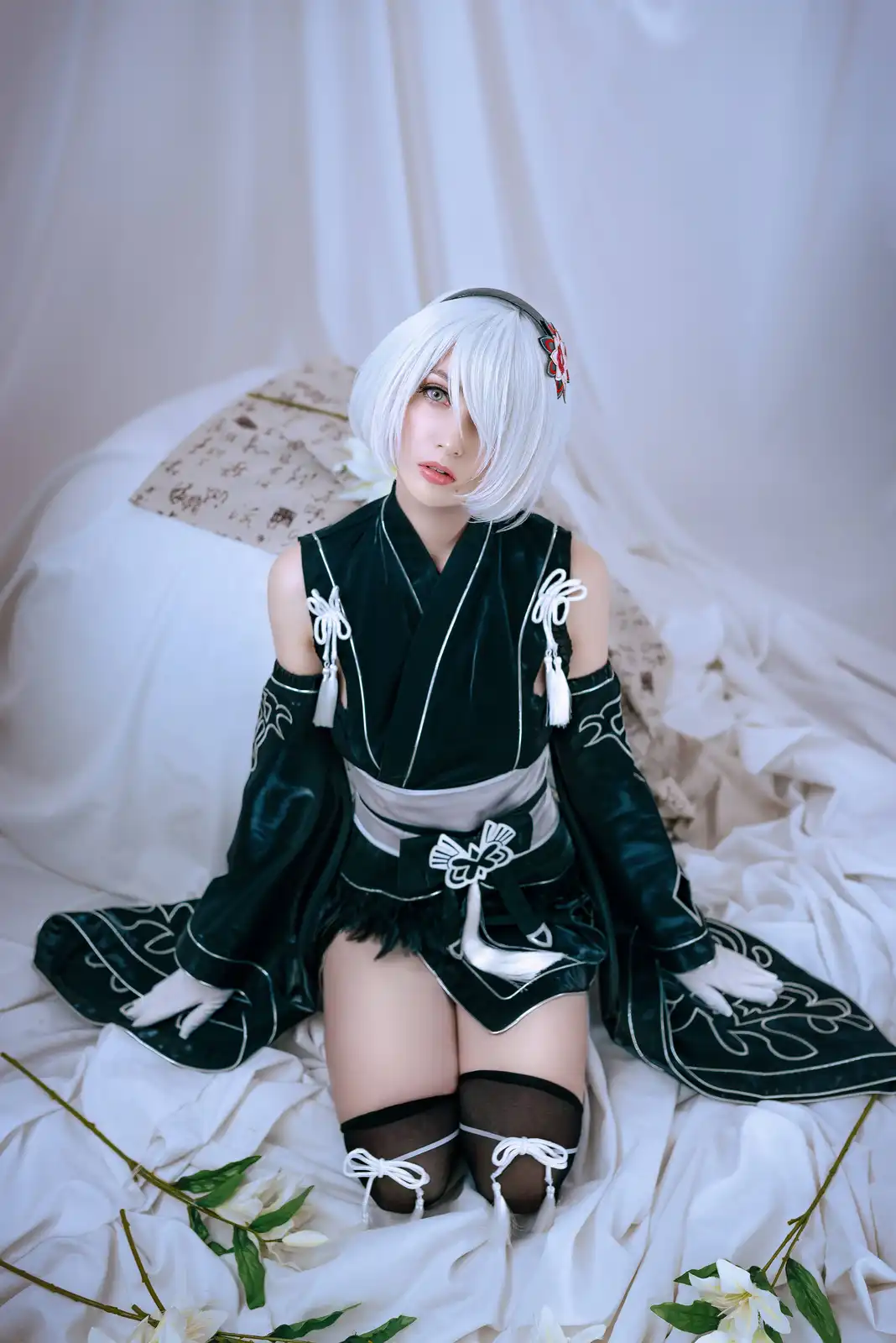A person in a detailed cosplay of a character with short white hair and a dark green, white-trimmed outfit sits on a white…