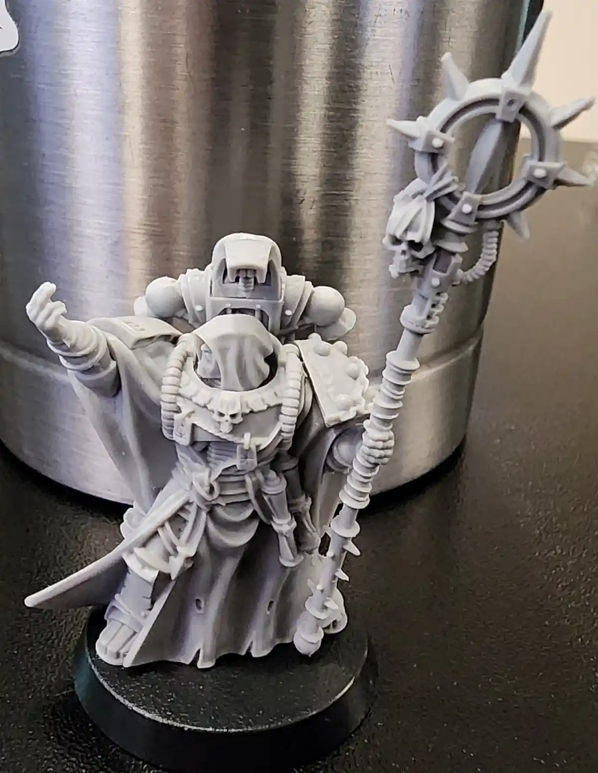 A gray, unpainted miniature of a fantasy warrior in ornate armor holding a spiked staff stands on a black base next to a…
