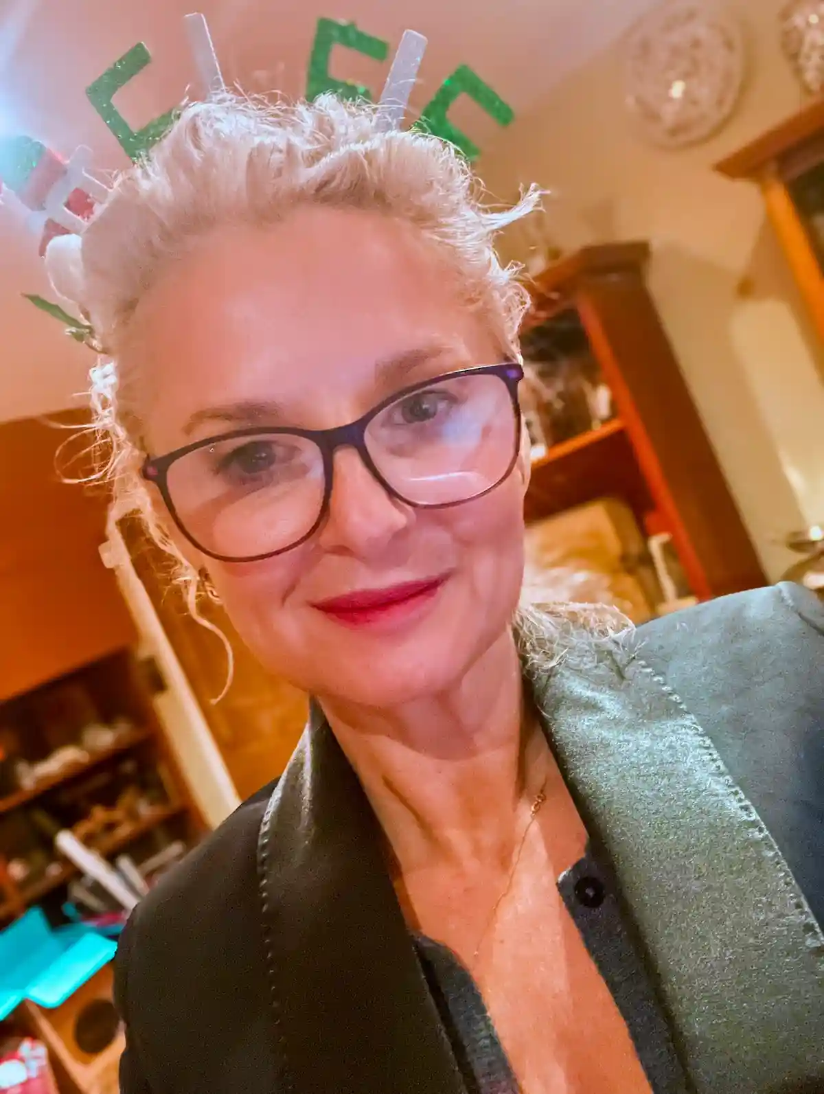A woman with short blonde hair and glasses smiles at the camera while wearing a green and white glittery headband shaped…
