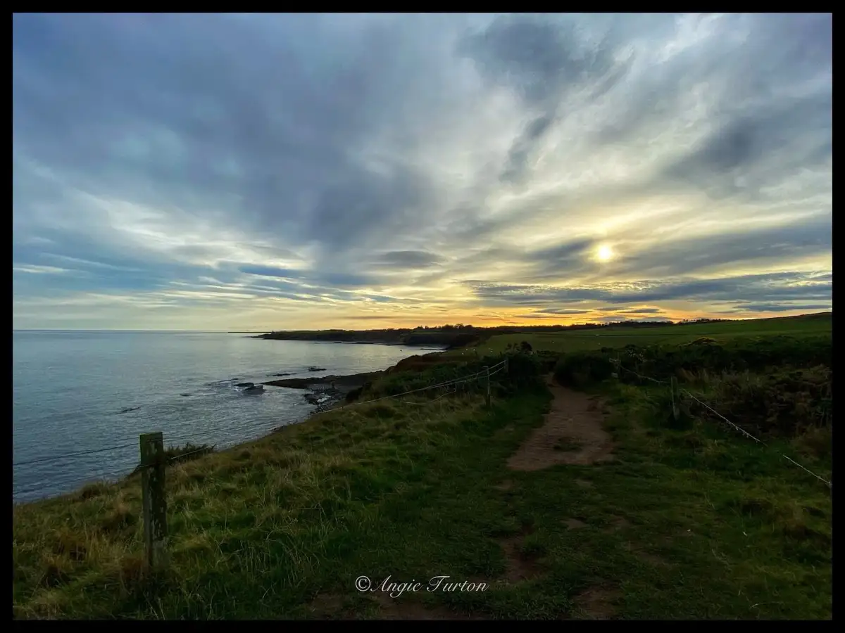 A scenic coastal path winds along a grassy cliff overlooking the ocean under a dramatic, cloudy sky with the sun low on the…