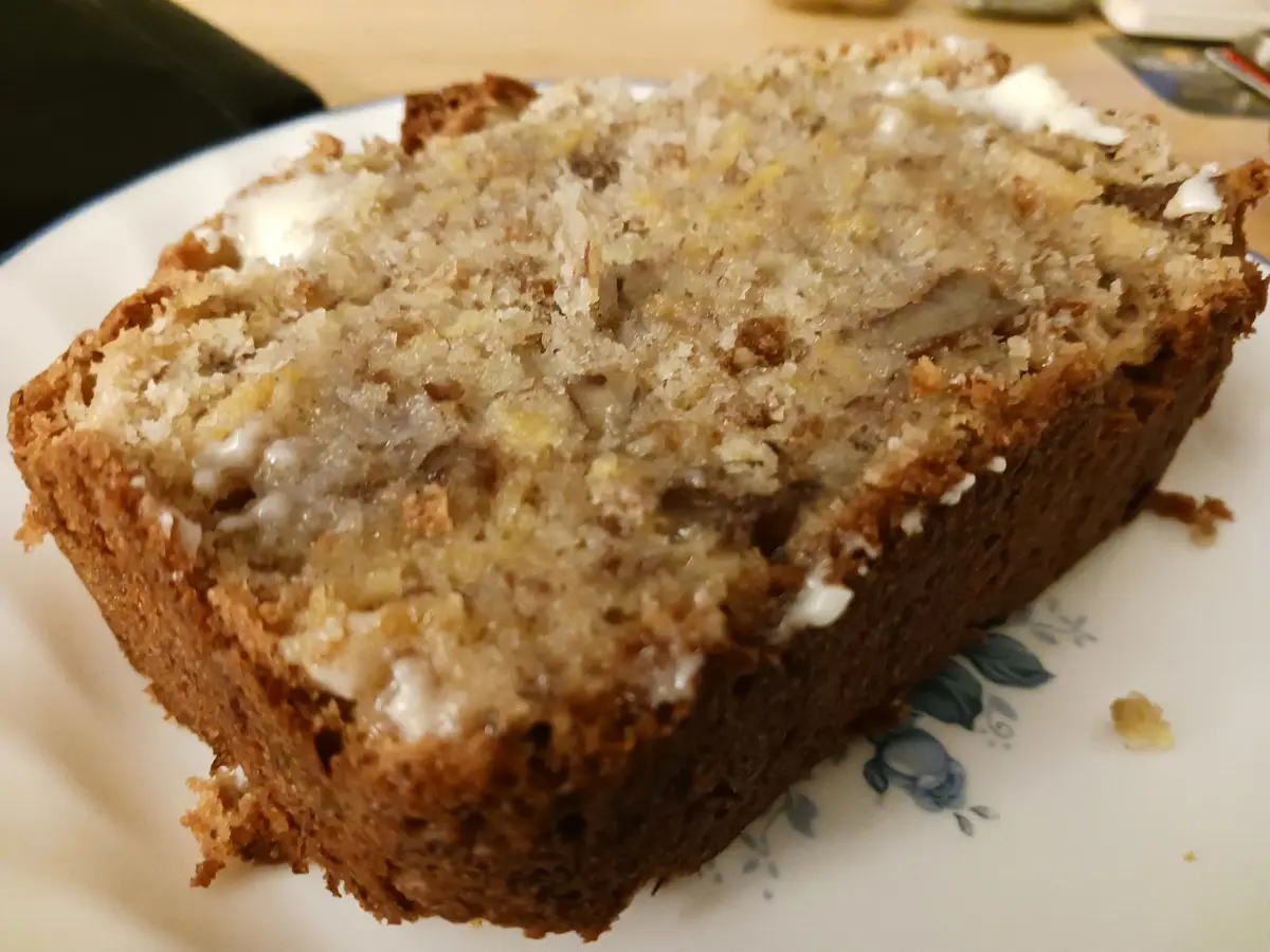 A close-up photo shows a thick slice of moist banana bread on a white plate with blue floral patterns, topped with a white…