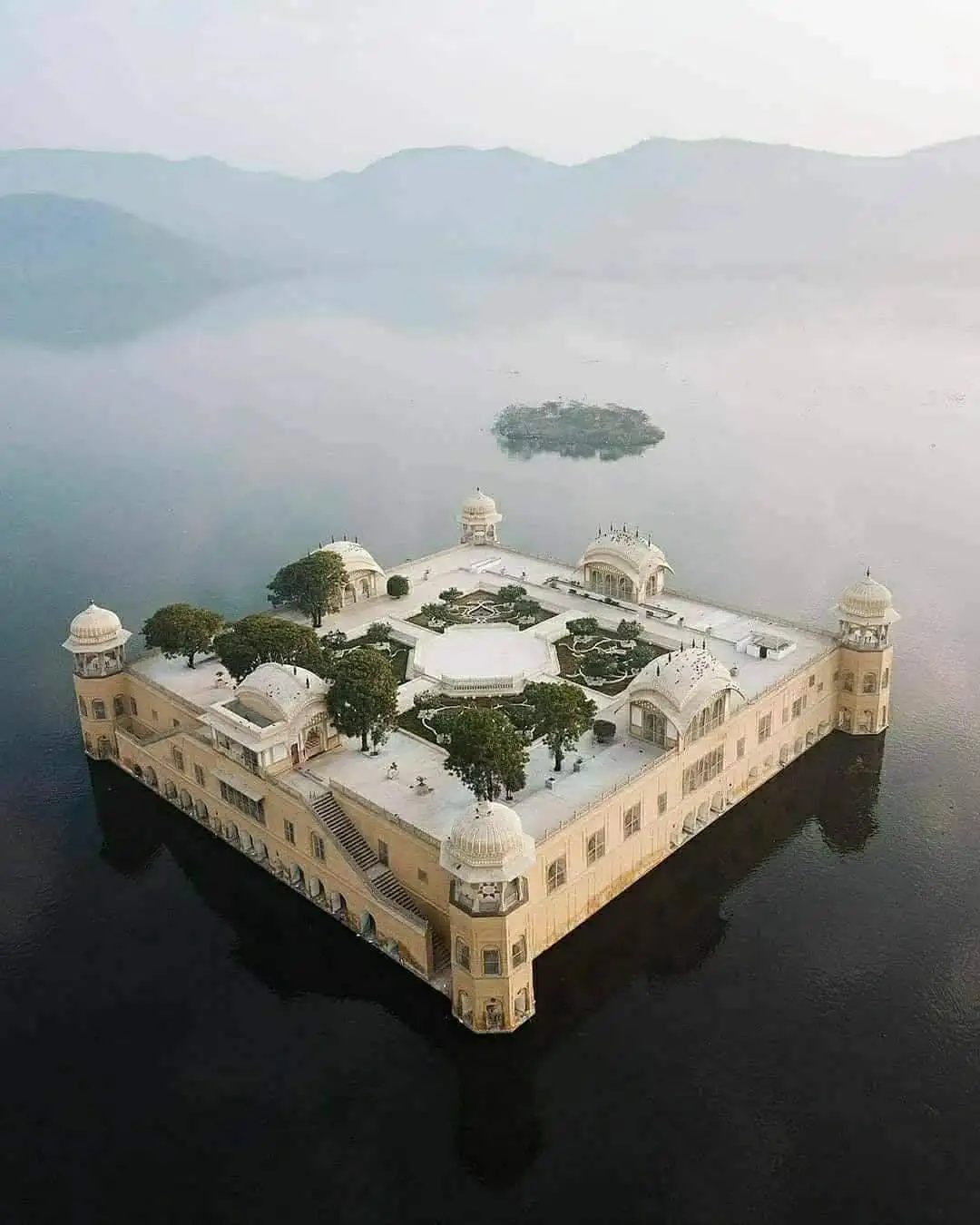 an aerial view shows the Lake Palace, a grand beige palace with domed towers and courtyards, floating on calm water with…