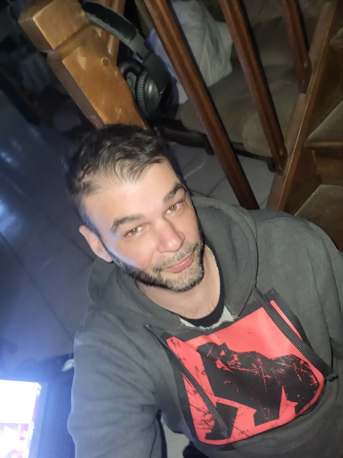 A man with a beard and short hair wearing a gray hoodie with a red and black graphic looks directly at the camera with a…