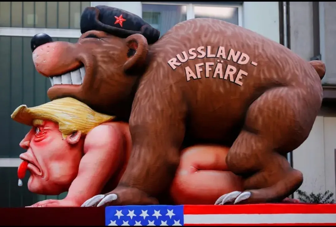 A large, cartoonish brown bear wearing a black military cap with a red star is depicted riding on top of a caricature of…