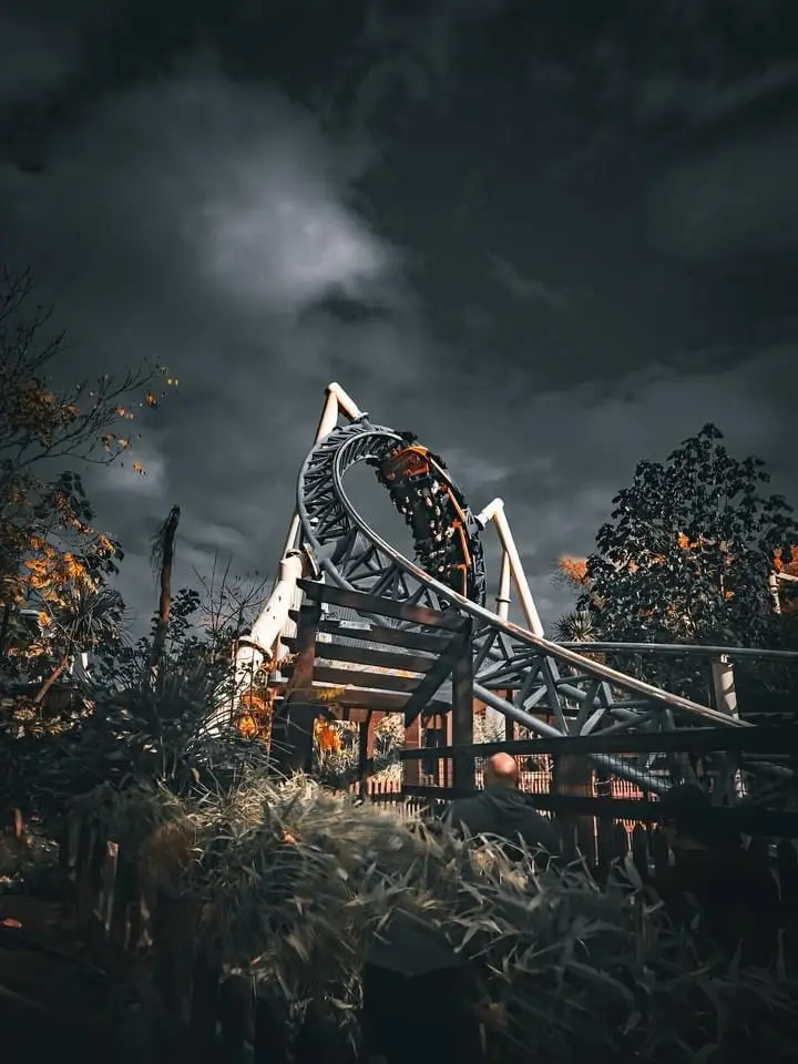 A dark, moody photo shows a roller coaster with a train at the top of a loop under a stormy sky, framed by trees and…