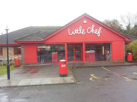 A red-brick Little Chef restaurant building with a sloped roof and large windows, featuring the brand's white cursive logo…