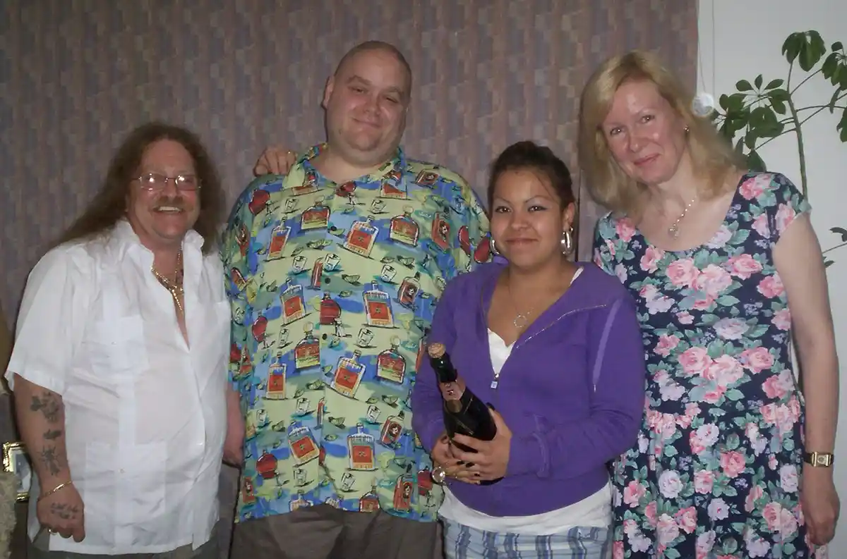 Four people pose for a photo indoors; a man in a white shirt with tattoos and glasses stands left, next to a bald man in a…