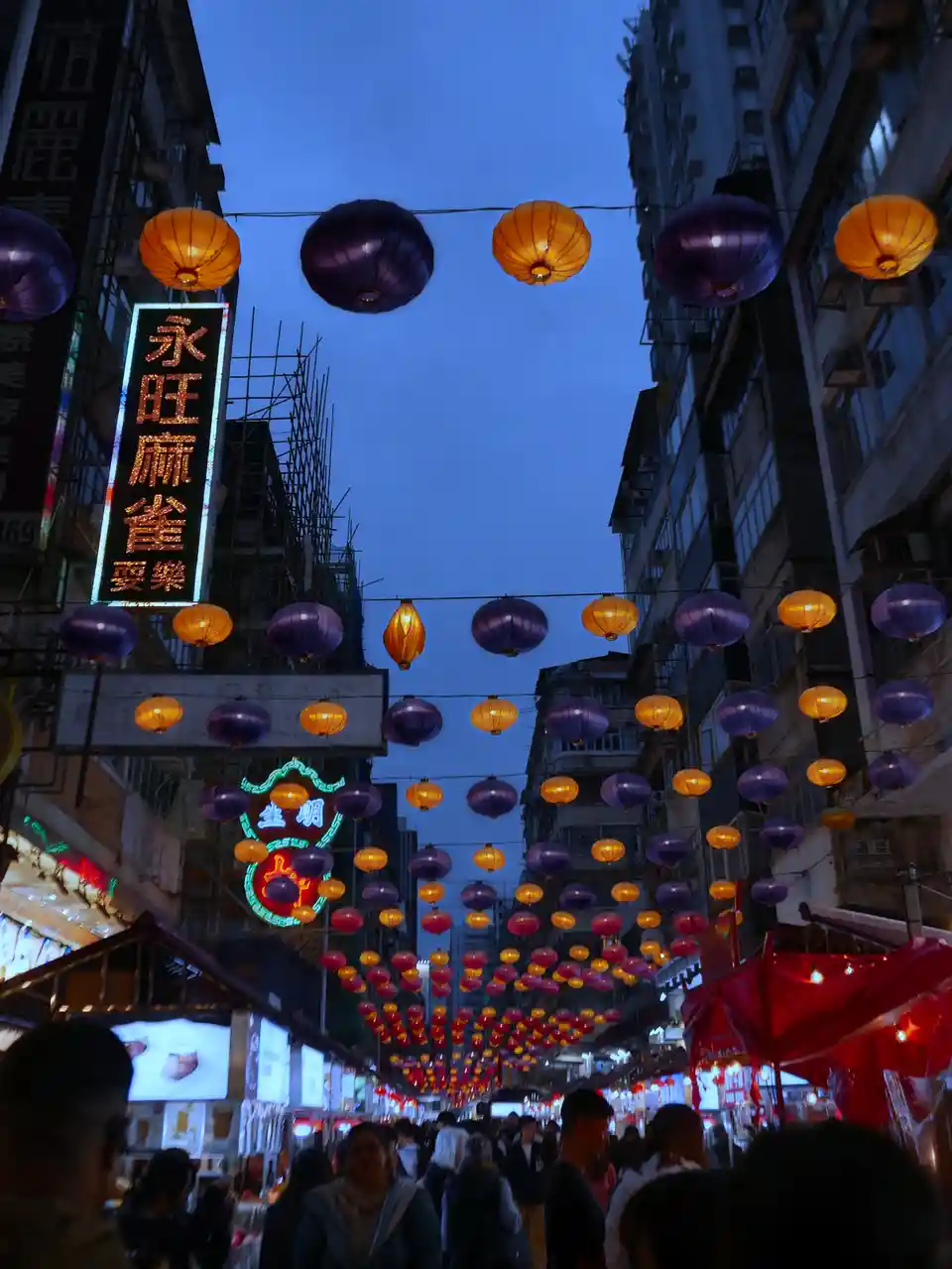 A bustling urban street at dusk is filled with crowds of people walking beneath strings of glowing orange and purple…