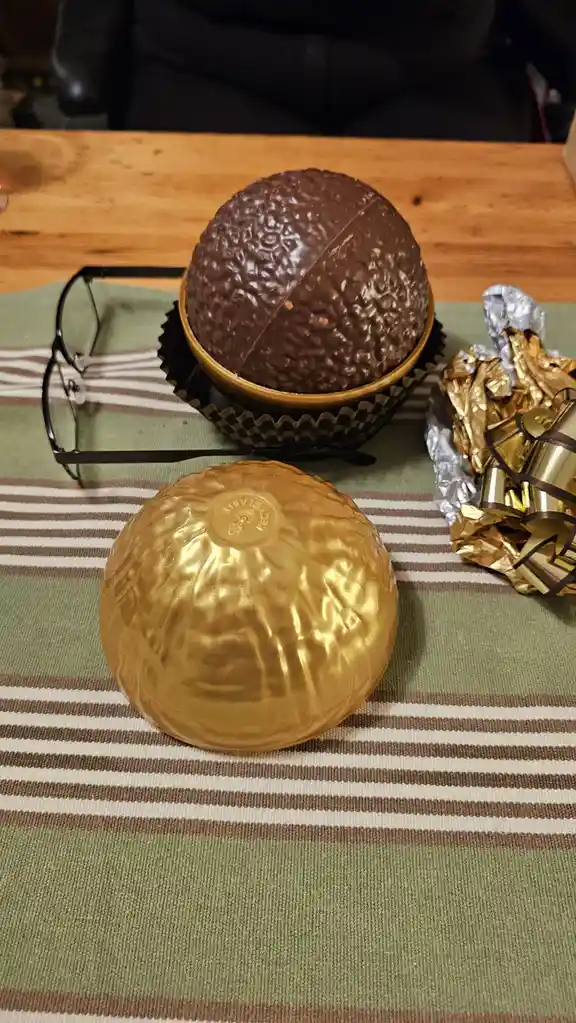 A large chocolate sphere sits in a cupcake liner on a striped placemat, next to a crumpled gold foil wrapper and a pair of…