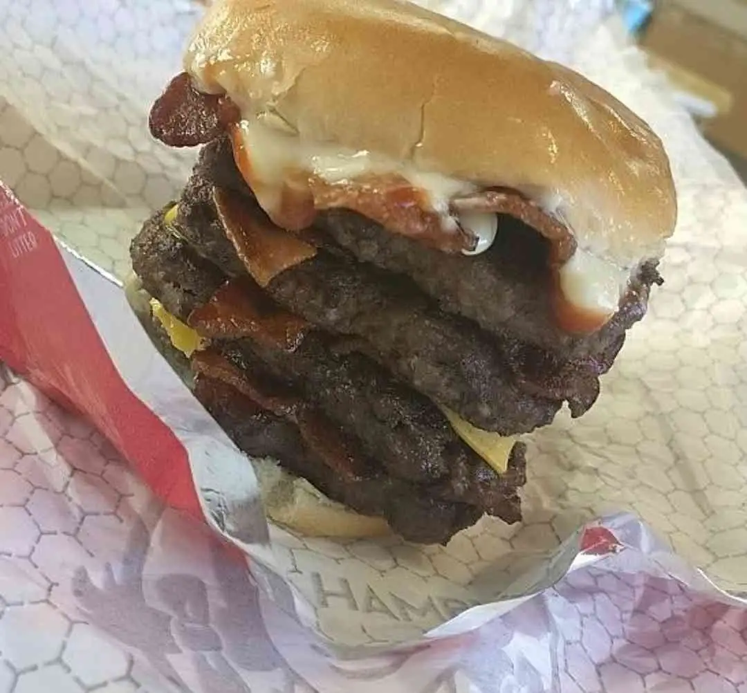 A towering, multi-patty burger with bacon and cheese oozing from between layers, wrapped in branded paper, with the text…