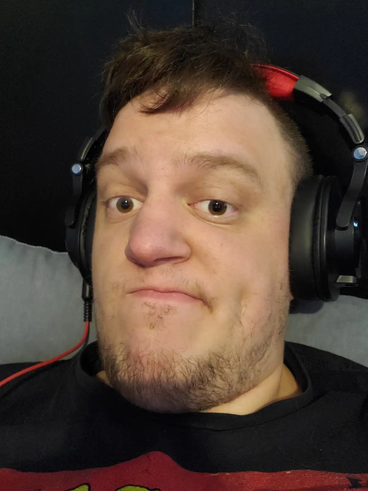 A man with brown hair and a beard, wearing black and red headphones, stares directly at the camera with wide eyes and a…