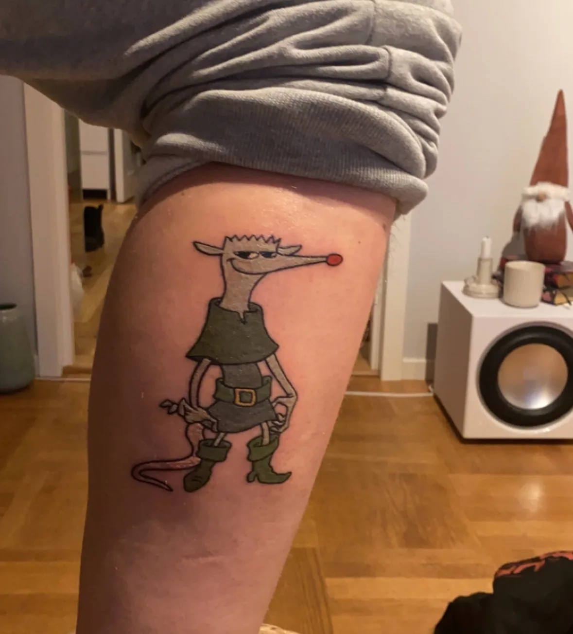 A person's leg is shown with a freshly inked tattoo of a cartoon rat character wearing green boots, a tunic, and a belt…