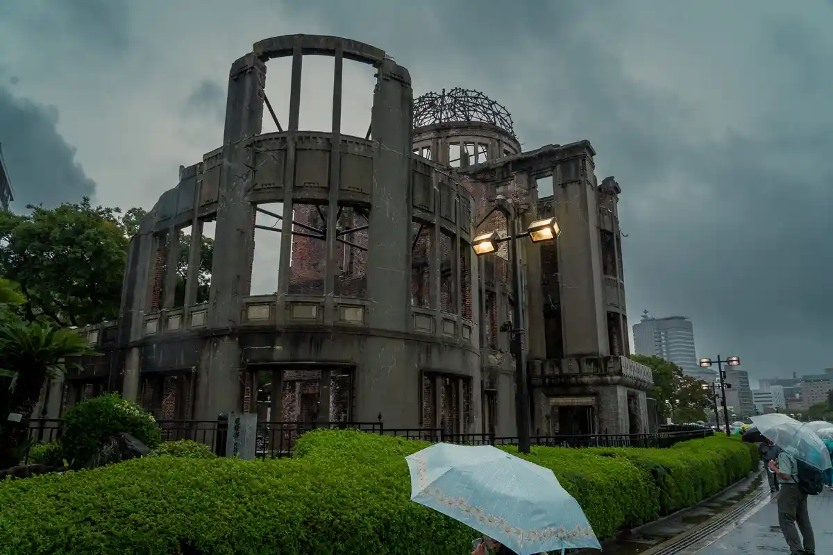The image shows the Hiroshima Peace Memorial (Genbaku Dome), a partially ruined building under a dark, rainy sky. In the…