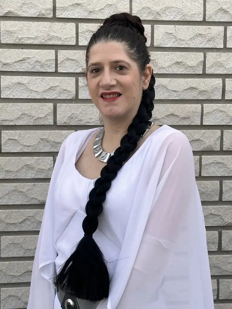 A woman with dark hair styled in a thick braid that ends in a black tassel, wearing a white sheer top and a silver necklace…