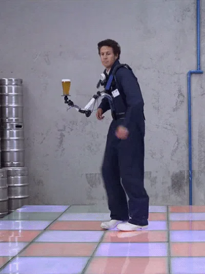 A man in a blue jumpsuit and harness stands on a colorful, illuminated dance floor, holding a beer on a mechanical arm. He…