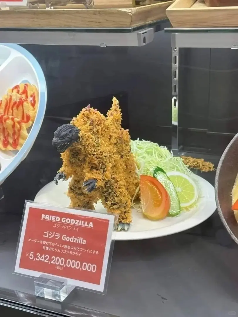 A large, crispy fried Godzilla-shaped food item sits on a white plate with garnishes, displayed behind glass. A red sign in…