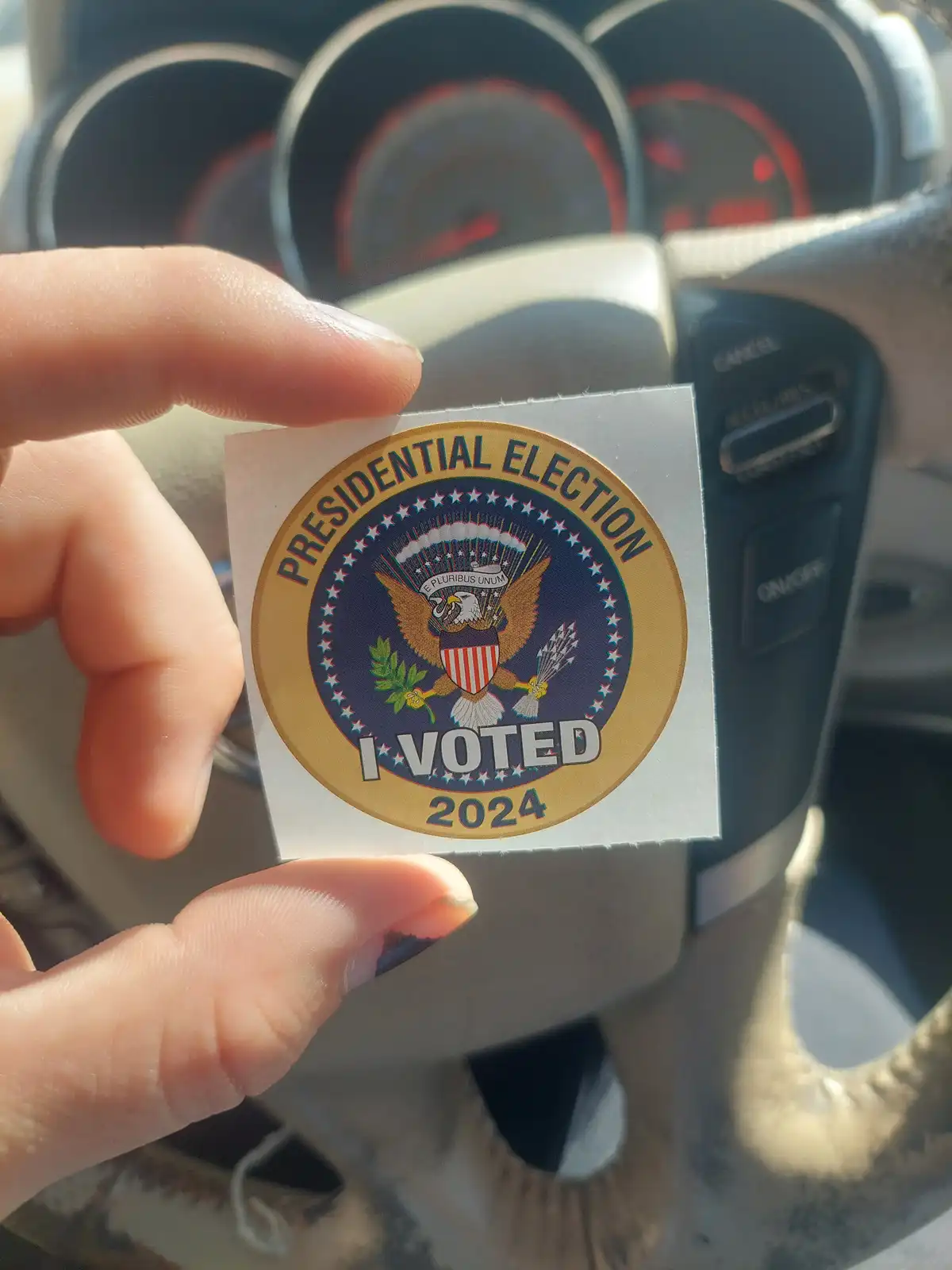 A hand holds up a circular 'I VOTED 2024' sticker featuring the presidential seal with an eagle, stars, and the motto 'E…