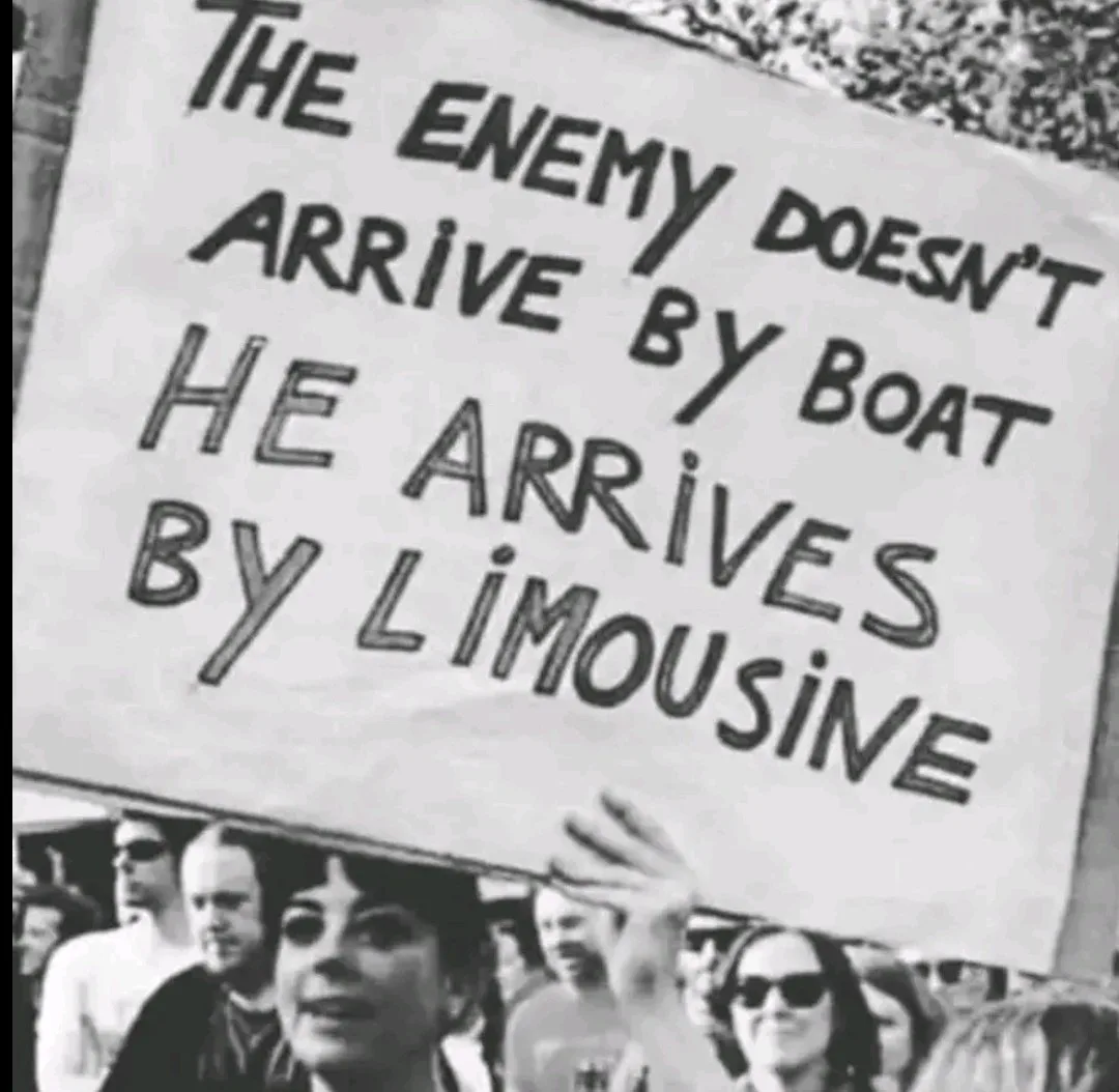A black-and-white photo shows a protest crowd with a person holding a handmade sign. The sign reads 'THE ENEMY DOESN'T…