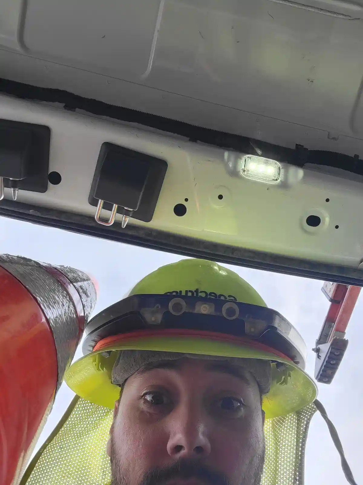 A low-angle selfie shows a man wearing a bright green hard hat with a black visor, looking directly at the camera with a…