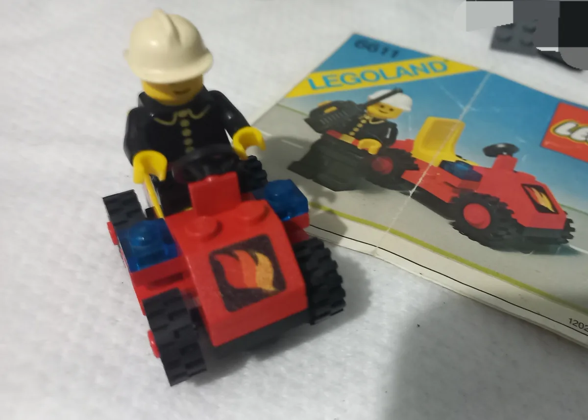 A close-up photo shows a Lego firefighter minifigure in a black uniform and white helmet, sitting in a red and black Lego…