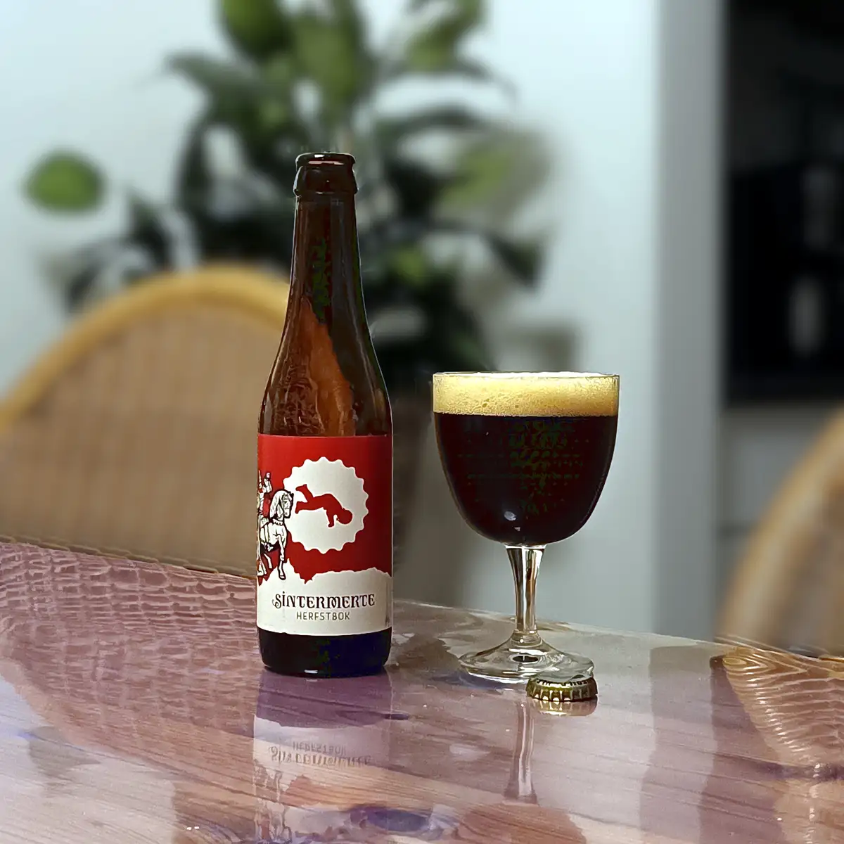 A brown beer bottle labeled 'Sintermerte Herfstbok' sits next to a stemmed glass filled with dark beer and a thick golden…