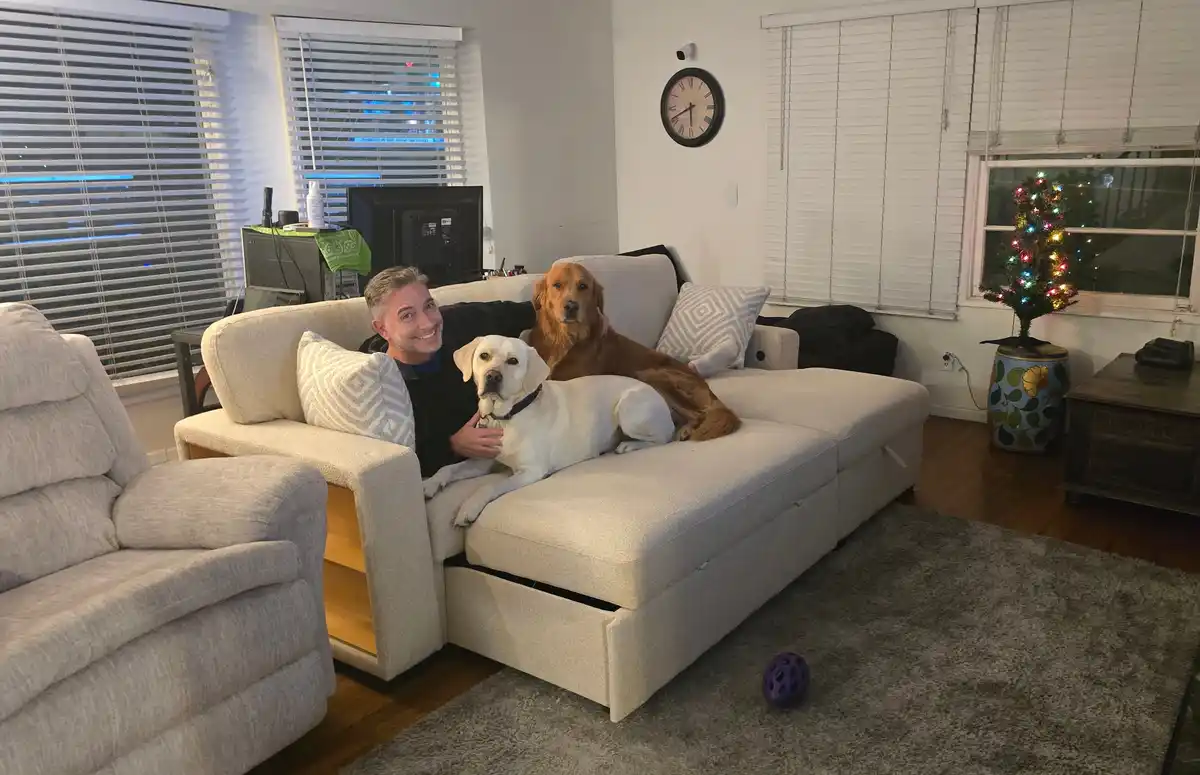 A man smiles at the camera while sitting on a beige couch with two dogs, a golden retriever and a yellow lab, beside him…