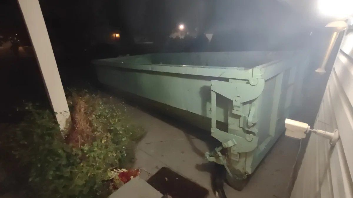 A nighttime photo shows a large, empty green dumpster placed beside a house, with a security camera mounted on the wall…