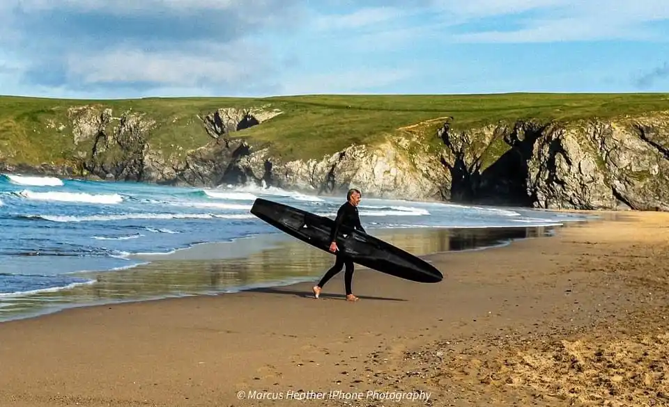 A person in a black wetsuit walks barefoot on a sandy beach carrying a black surfboard under their arm. Behind them, waves…