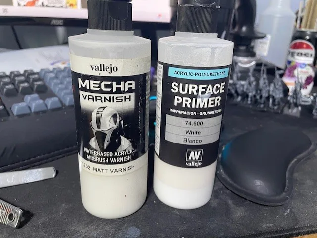 Two white paint bottles from Vallejo sit side by side on a desk next to a keyboard and mousepad. The left bottle is labeled…
