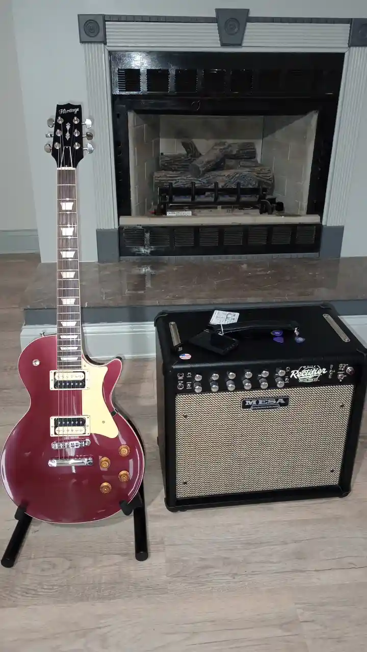 A red electric guitar with a cream pickguard and gold hardware stands on a stand next to a black Mesa Rectifier guitar…