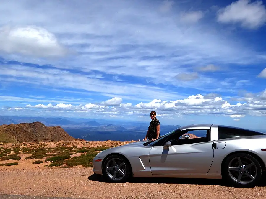 A person wearing sunglasses and a dark patterned shirt stands beside a silver Chevrolet Corvette parked on a dirt overlook…