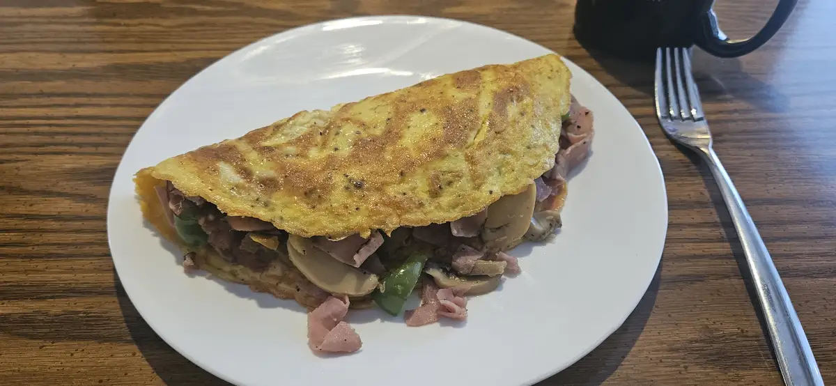 A close-up photo of a folded omelette filled with ham, mushrooms, and green peppers on a white plate, set on a wooden table…