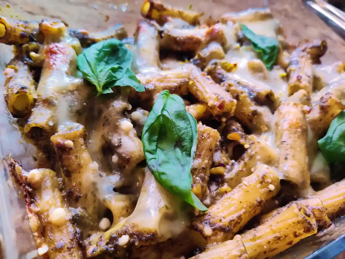 A close-up photo of baked pasta with melted cheese, pesto sauce, pine nuts, and fresh basil leaves in a glass baking dish…