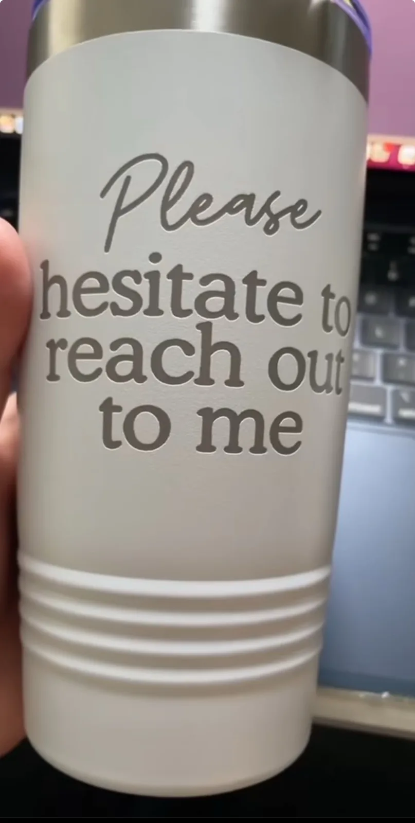 A close-up photo shows a hand holding a white, frosted stainless steel tumbler with a silver lid. The tumbler has engraved…