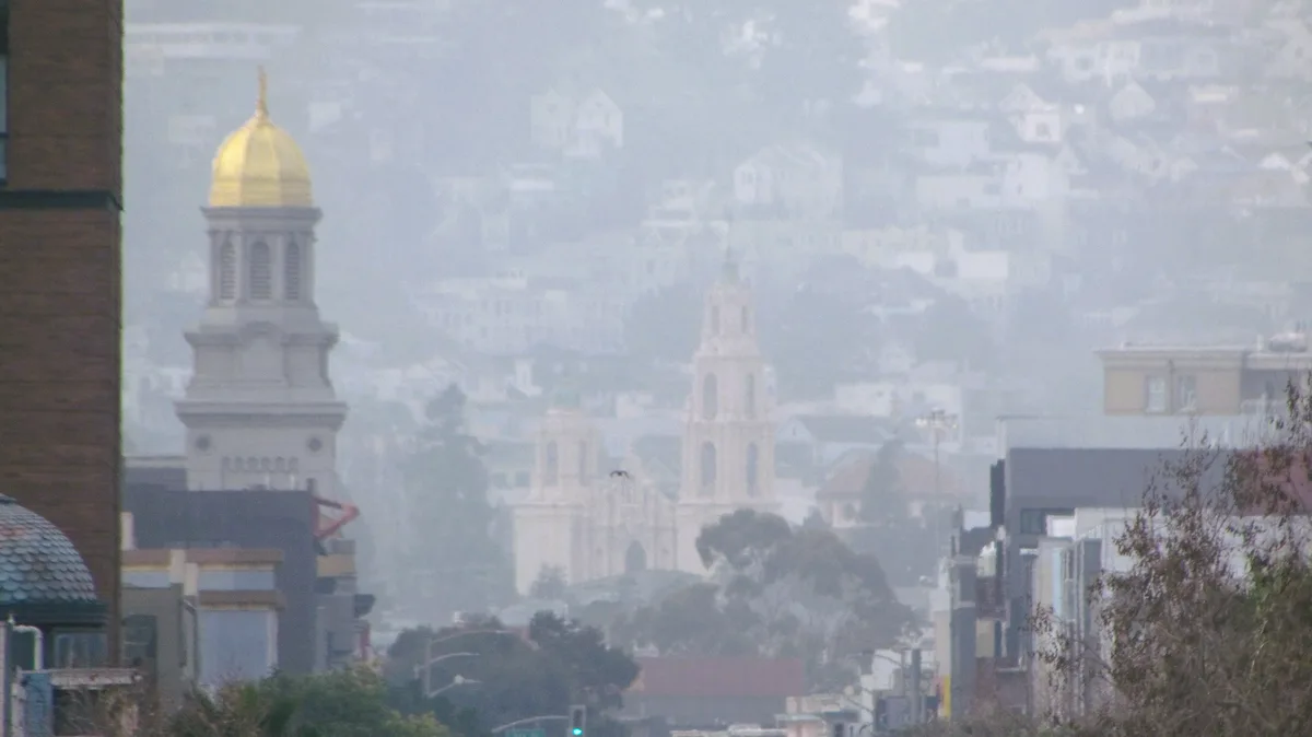 A hazy cityscape with a prominent building featuring a golden dome in the foreground, with other buildings and hills visible…