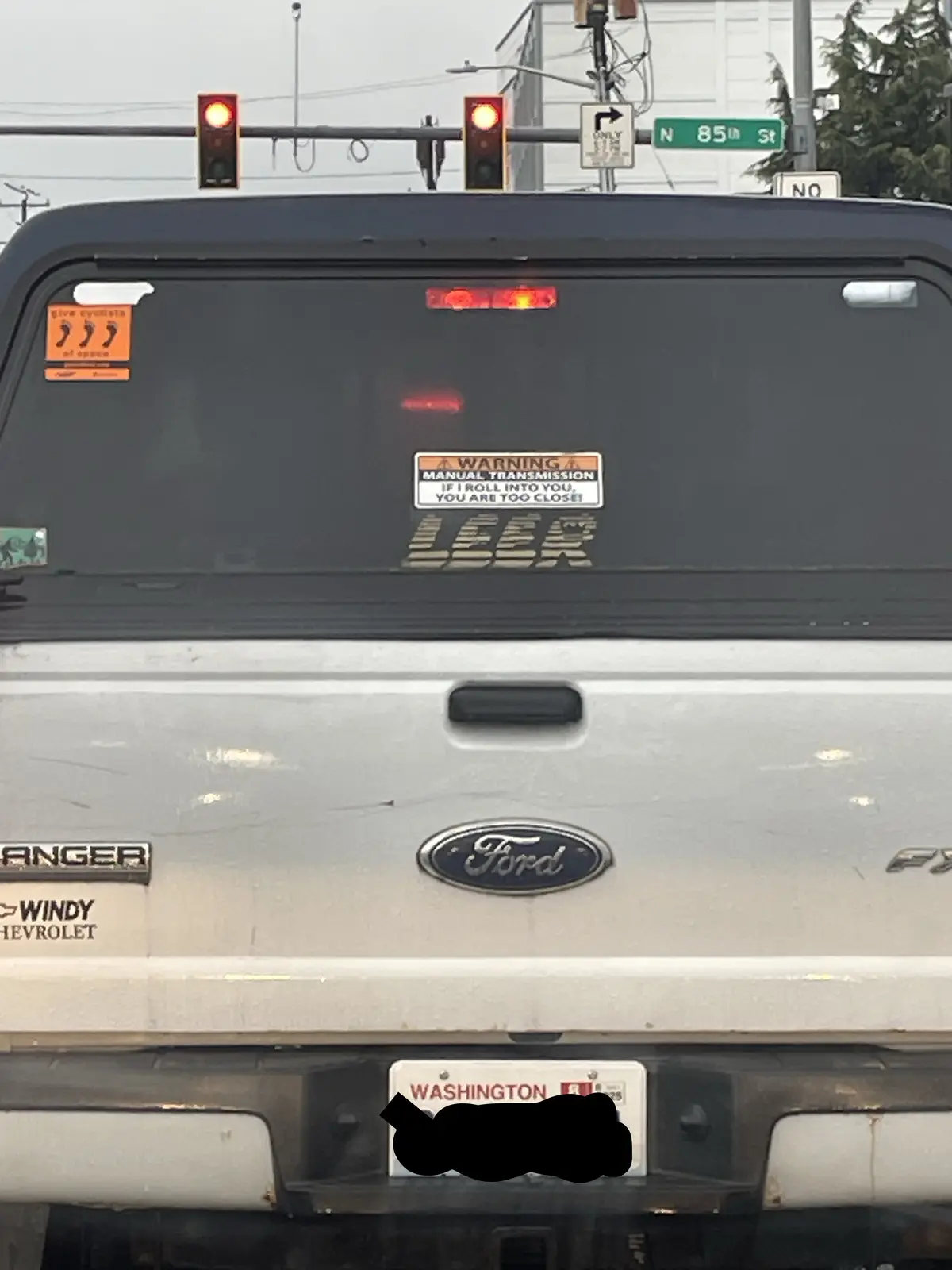 The rear of a silver Ford Ranger pickup truck is shown stopped at a red light, with a prominent warning sticker on the back…