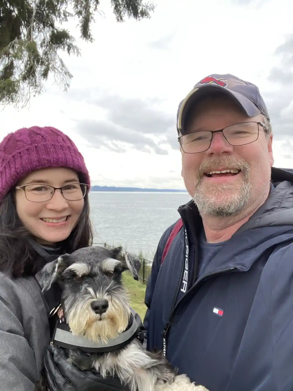 A selfie of a smiling man and woman outdoors near a body of water, holding a small black-and-silver schnauzer. The woman…
