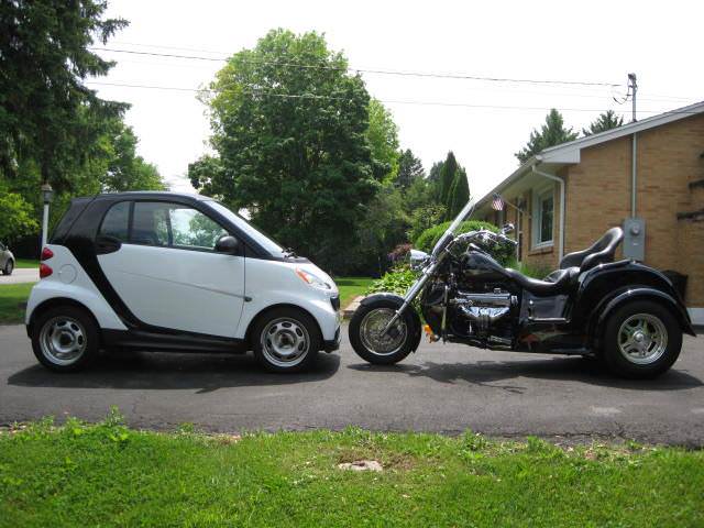 A white and black smart car is parked side-by-side with a large black three-wheeled motorcycle on a residential street. The…