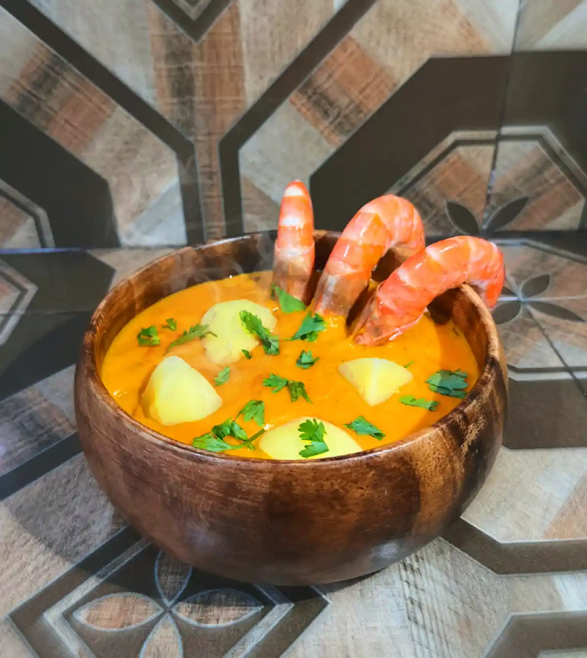 A wooden bowl filled with orange-colored soup, garnished with chopped green herbs and chunks of potato, with three cooked…