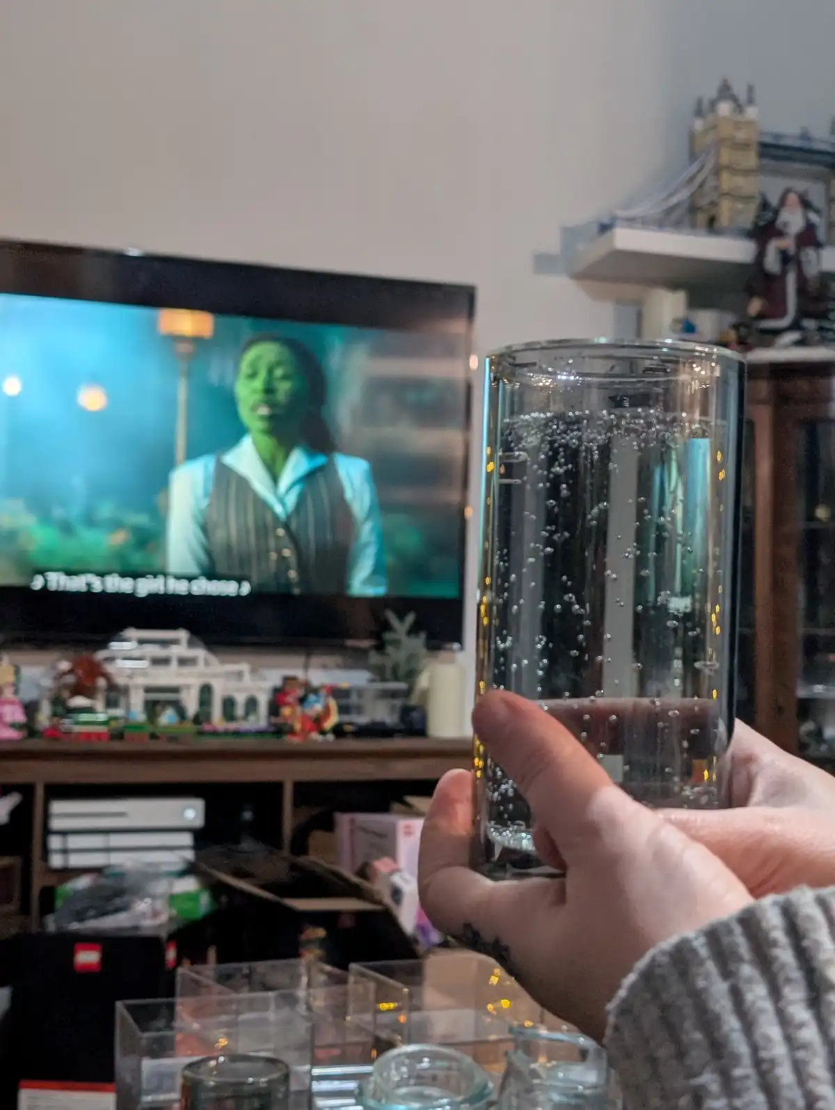 A person holds a tall glass of sparkling water in front of a TV showing a green-skinned woman singing, with the subtitle…