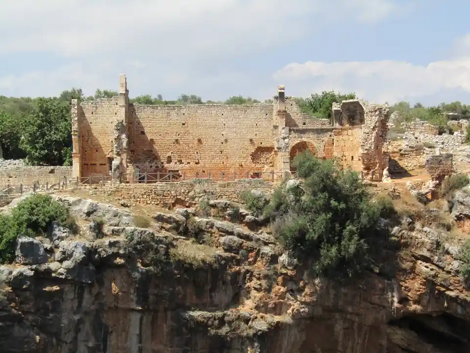 The image shows ancient stone ruins perched on a steep, rocky cliffside under a partly cloudy sky. A person in a…