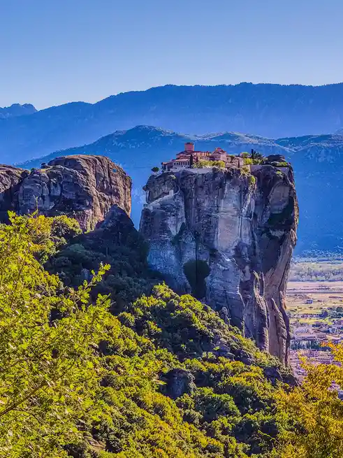 A scenic photo of a monastery perched atop a towering, sheer rock pillar in a mountainous landscape, with lush green trees…