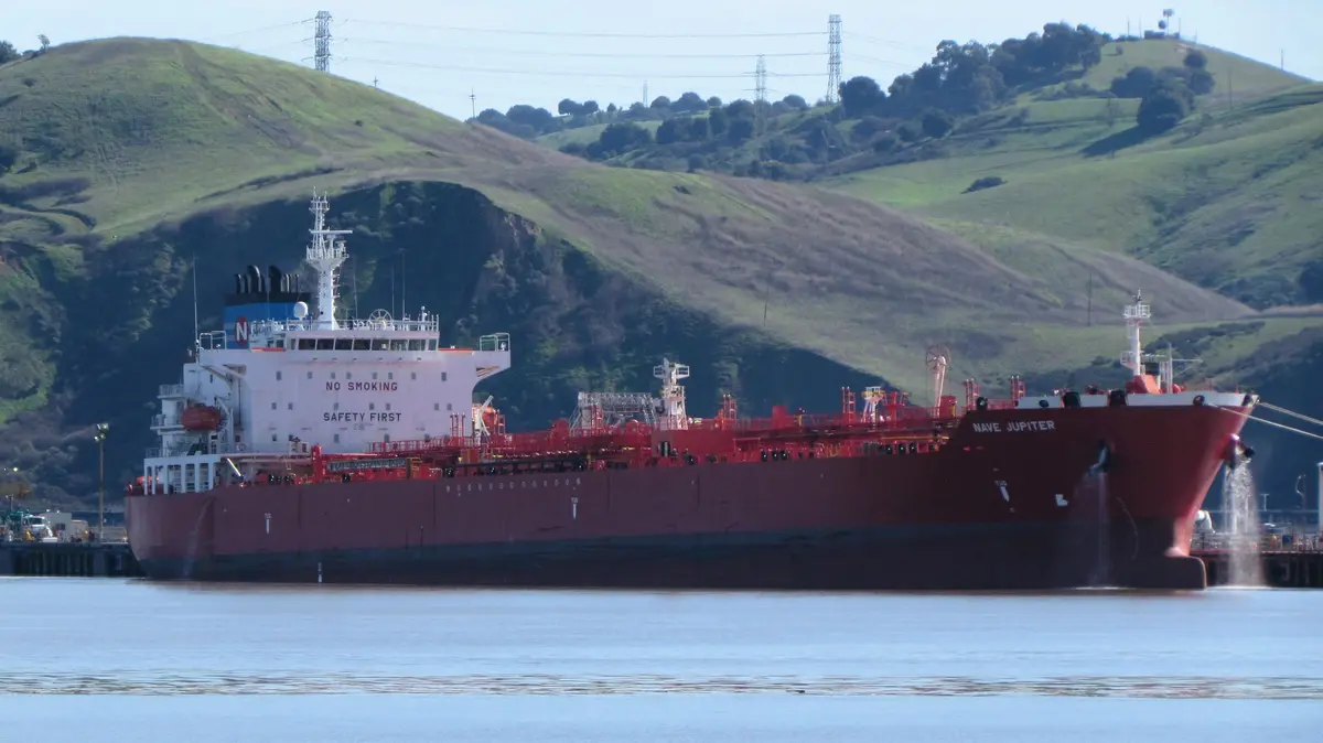 A large red and white cargo ship named 'NAVE JUPITER' is docked in water with green hills and power lines in the background…