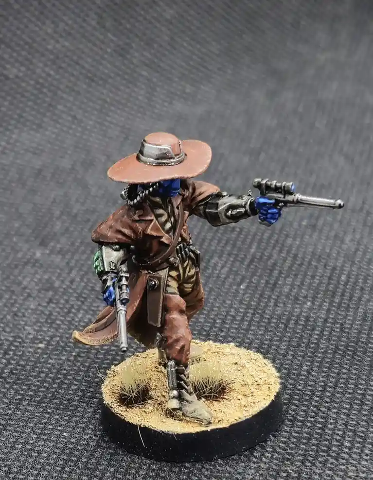 A painted miniature figure of a space cowboy wearing a brown hat, blue face covering, and brown coat, standing on a…