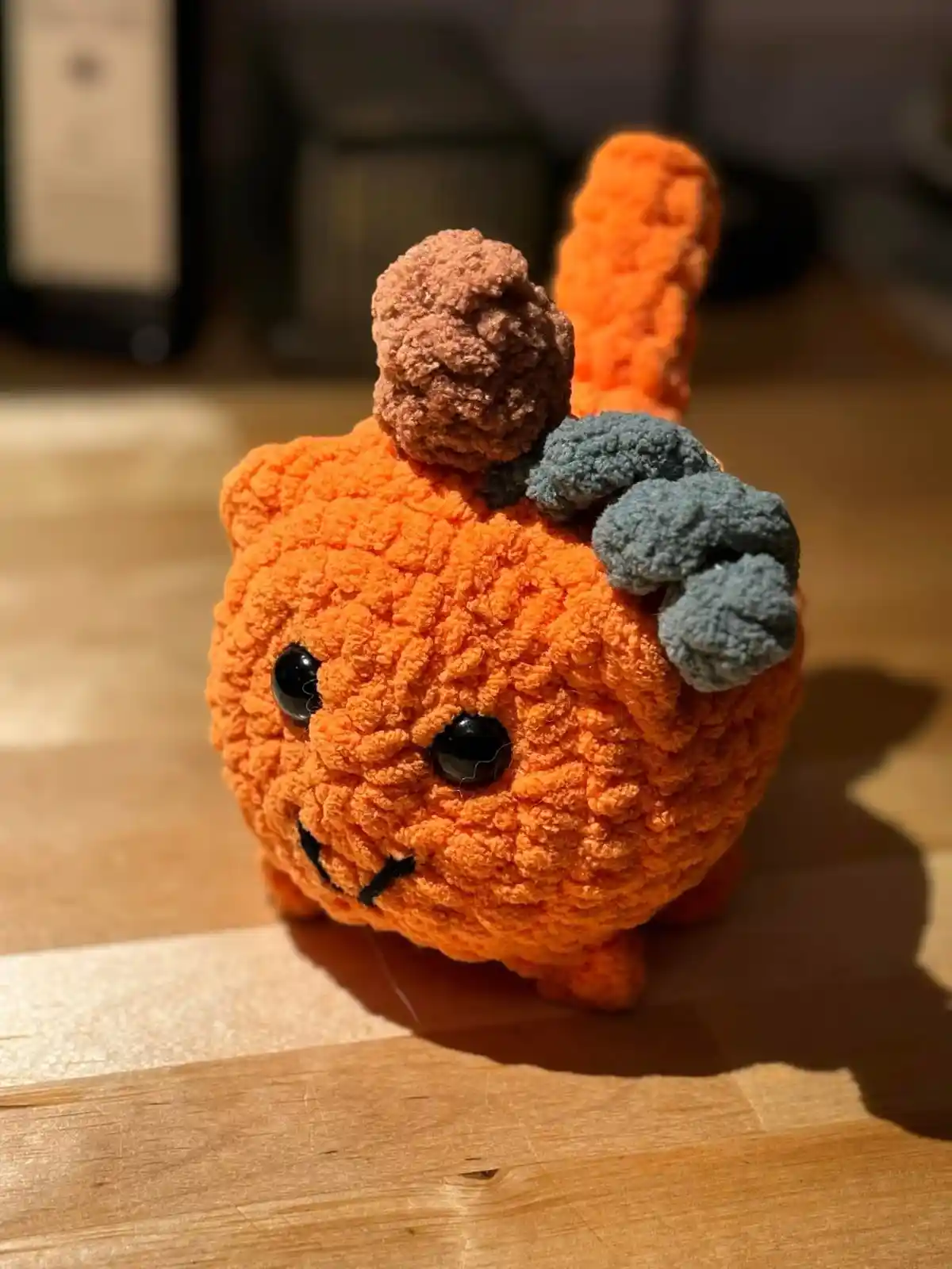 A close-up photo of a handmade, orange, textured crochet cat figurine with a brown pom-pom on its head and grey yarn ears…