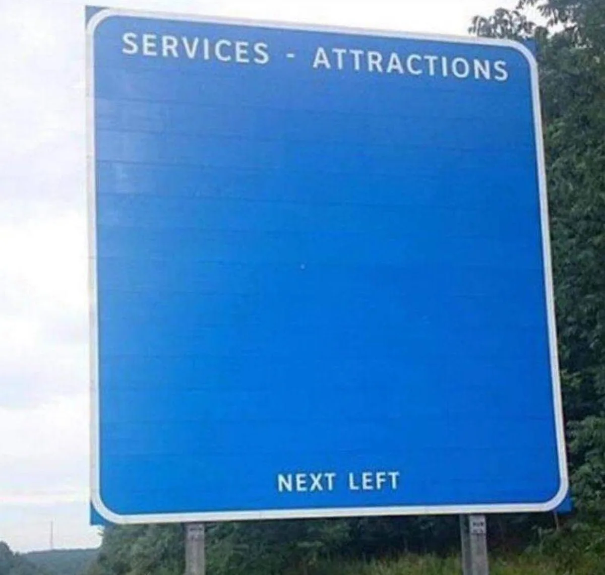 A large blue highway sign with white text reading 'SERVICES - ATTRACTIONS' at the top and 'NEXT LEFT' at the bottom, but the…