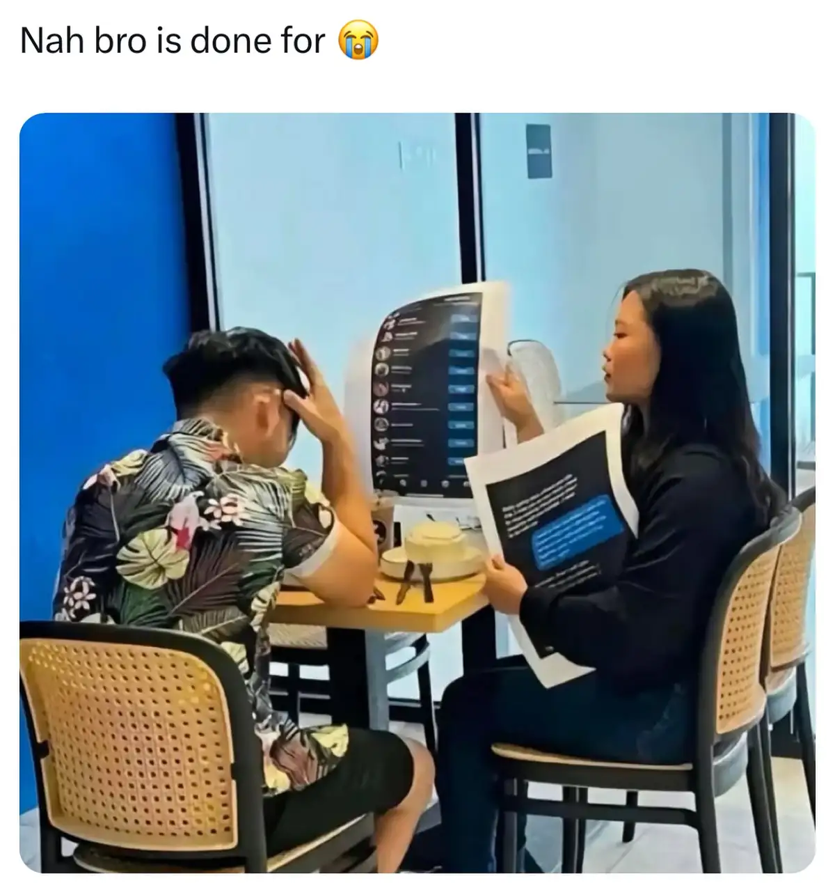 A man in a floral shirt sits at a restaurant table, hand on his head in despair, while a woman in a black shirt holds up a…