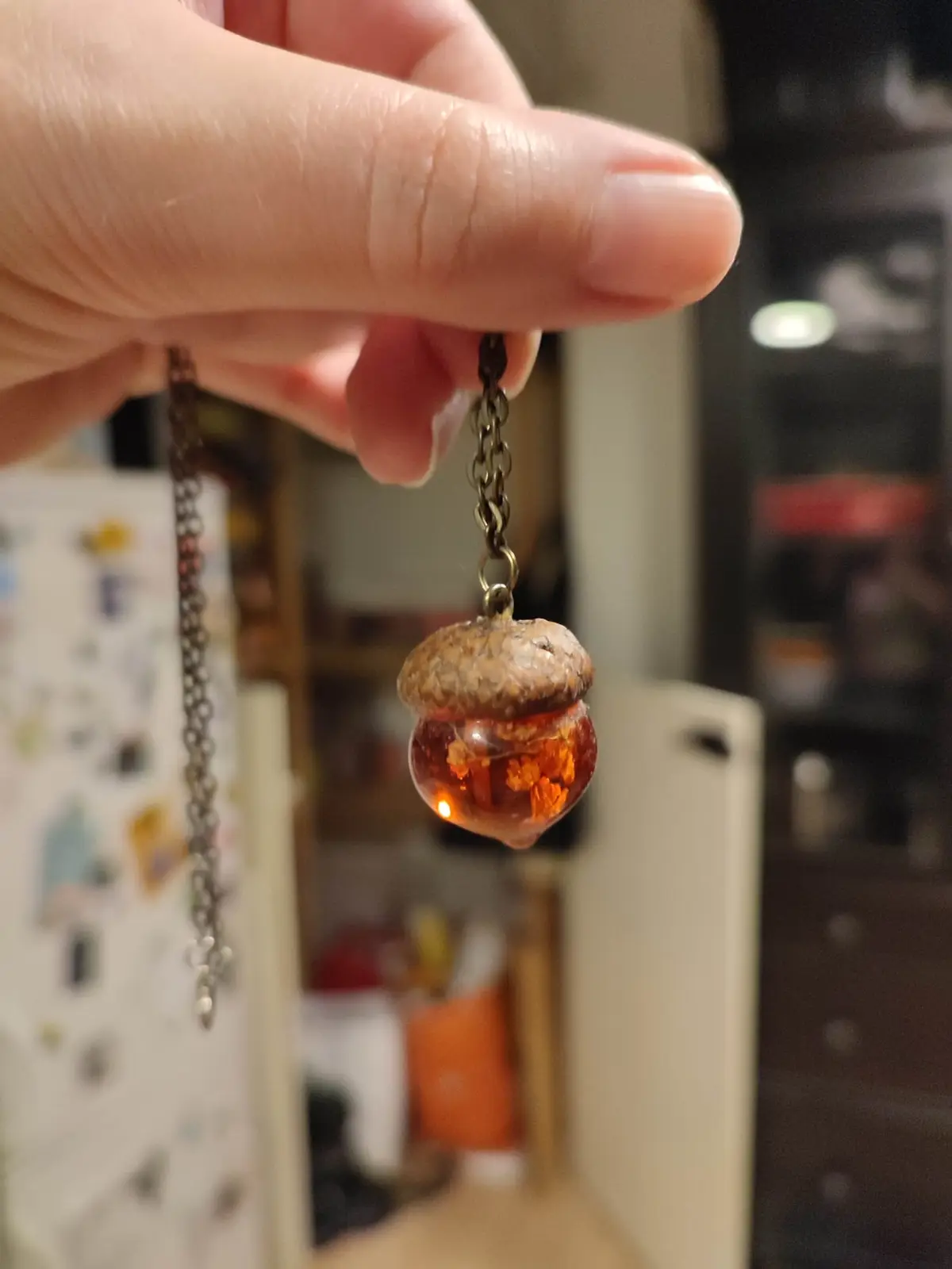 A hand holds a necklace pendant shaped like an acorn, with a translucent amber-colored lower half containing tiny orange…
